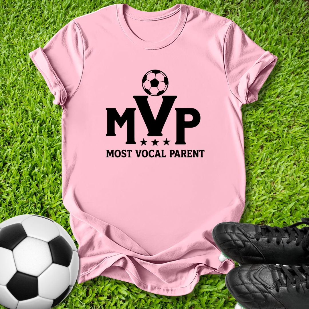 A light pink short-sleeve t-shirt laid flat on green grass, featuring a black graphic that reads 'MVP MOST VOCAL PARENT' with a soccer ball above the letters. A soccer ball and black cleats are placed around it as props.
