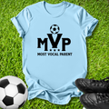 A light blue short-sleeve t-shirt laid flat on green grass, featuring a black graphic that reads 'MVP MOST VOCAL PARENT' with a soccer ball above the letters. A soccer ball and black cleats are placed around it as props.