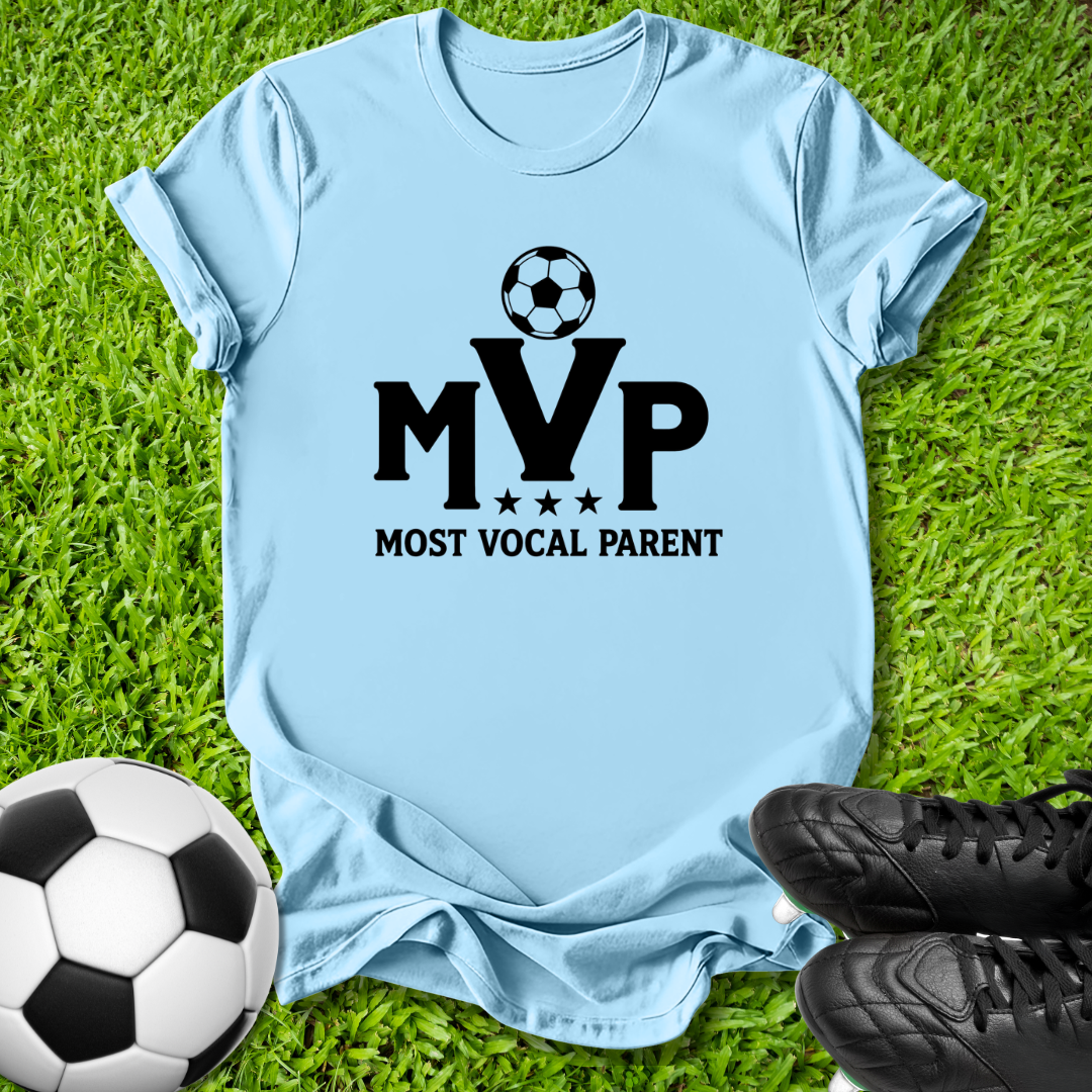 A light blue short-sleeve t-shirt laid flat on green grass, featuring a black graphic that reads 'MVP MOST VOCAL PARENT' with a soccer ball above the letters. A soccer ball and black cleats are placed around it as props.