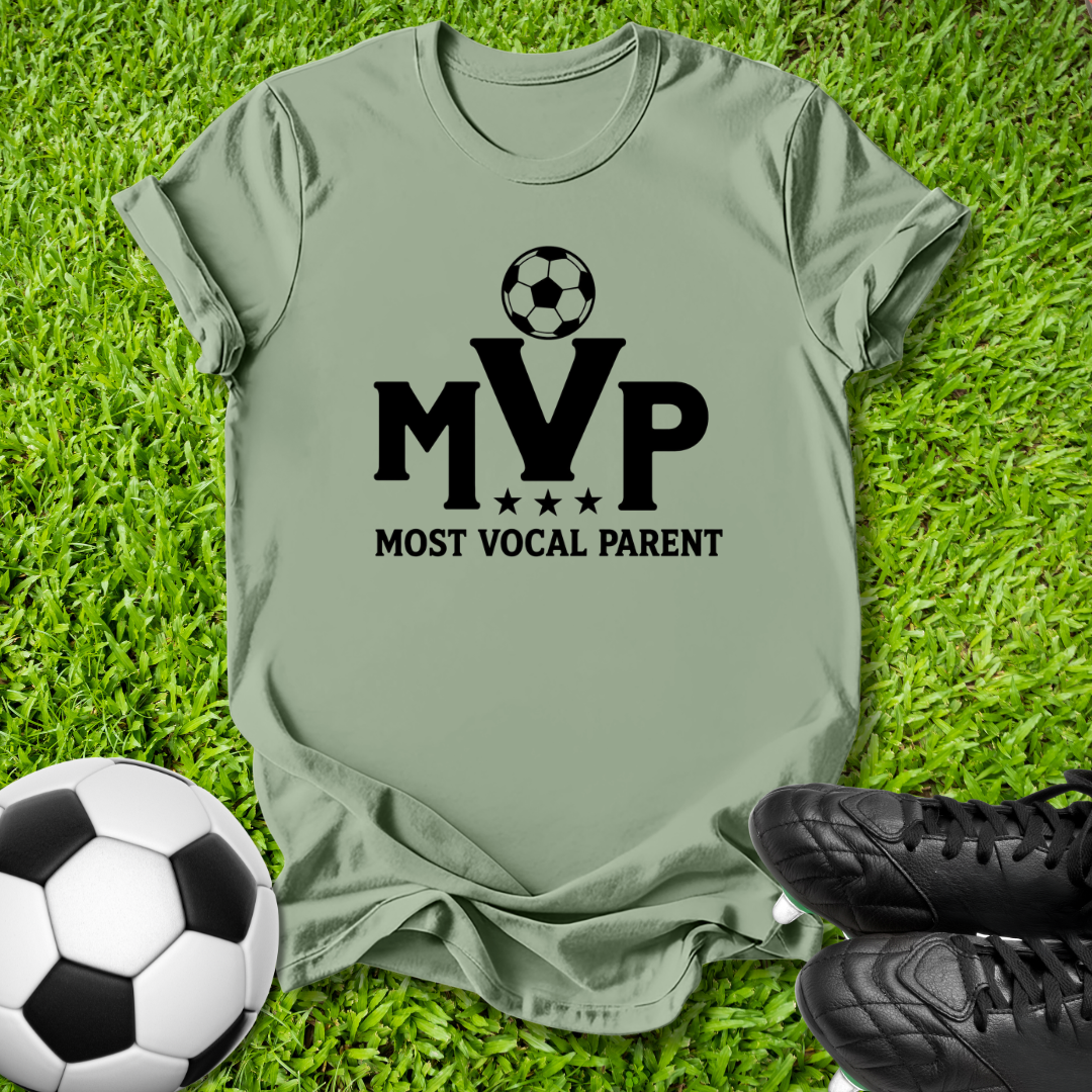 Green short-sleeve T-shirt laid flat on green grass, featuring a black soccer ball graphic above the letters 'MVP' with 'MOST VOCAL PARENT' underneath. A soccer ball and black cleats are placed around it as props.