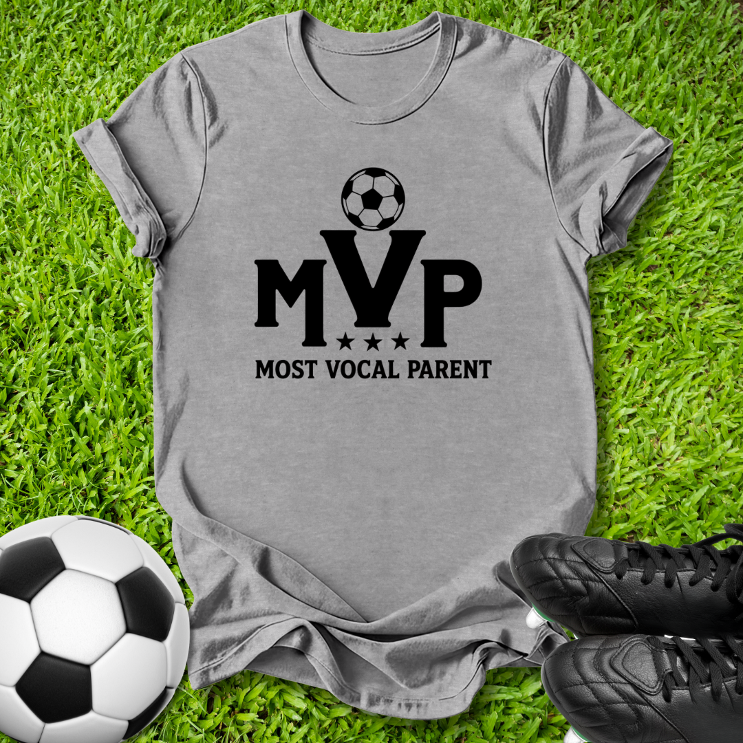 A heather gray short-sleeve t-shirt with a black graphic reading 'MVP MOST VOCAL PARENT' and a soccer ball icon, laid flat on green grass with a soccer ball and black cleats nearby.