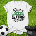 A white graphic t-shirt laid flat on green grass, featuring the text 'Best SOCCER GRANDMA Ever!' in black and green lettering with a soccer ball icon. A soccer ball and black cleats are placed around it as props.