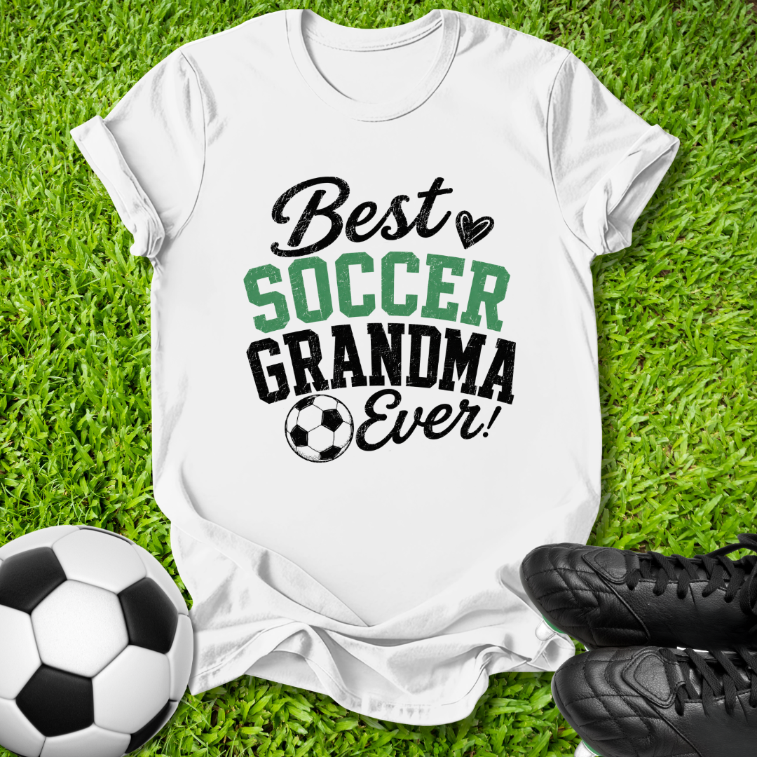 A white graphic t-shirt laid flat on green grass, featuring the text 'Best SOCCER GRANDMA Ever!' in black and green lettering with a soccer ball icon. A soccer ball and black cleats are placed around it as props.