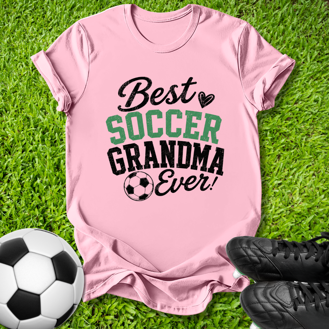 A pink short-sleeve graphic T-shirt laid flat on green grass, with a soccer ball on the left and black cleats on the right. The shirt reads 'Best SOCCER GRANDMA Ever!' in black and green lettering with a small soccer ball graphic.