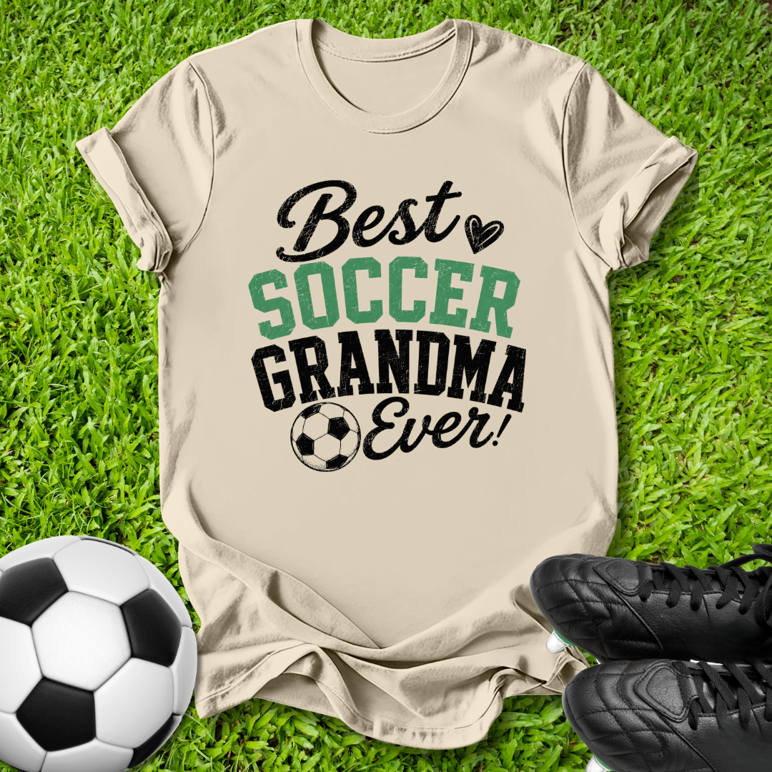 A beige graphic t-shirt laid flat on green grass, featuring the text 'Best SOCCER GRANDMA Ever!' in black and green lettering with a soccer ball graphic. A soccer ball and black cleats are placed around it as props.