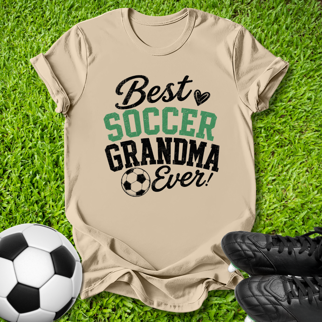 A beige graphic t-shirt laid flat on green grass, featuring the text 'Best SOCCER GRANDMA Ever!' in black and green lettering with a soccer ball icon. A soccer ball and black cleats are placed around it as props.