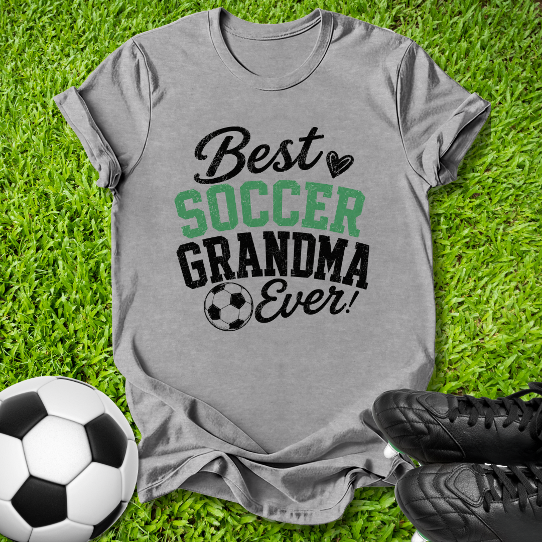 A heather gray graphic t-shirt laid flat on green grass, featuring the text 'Best SOCCER GRANDMA Ever!' in black and green lettering with a soccer ball icon. A soccer ball and black cleats are placed around it as props.