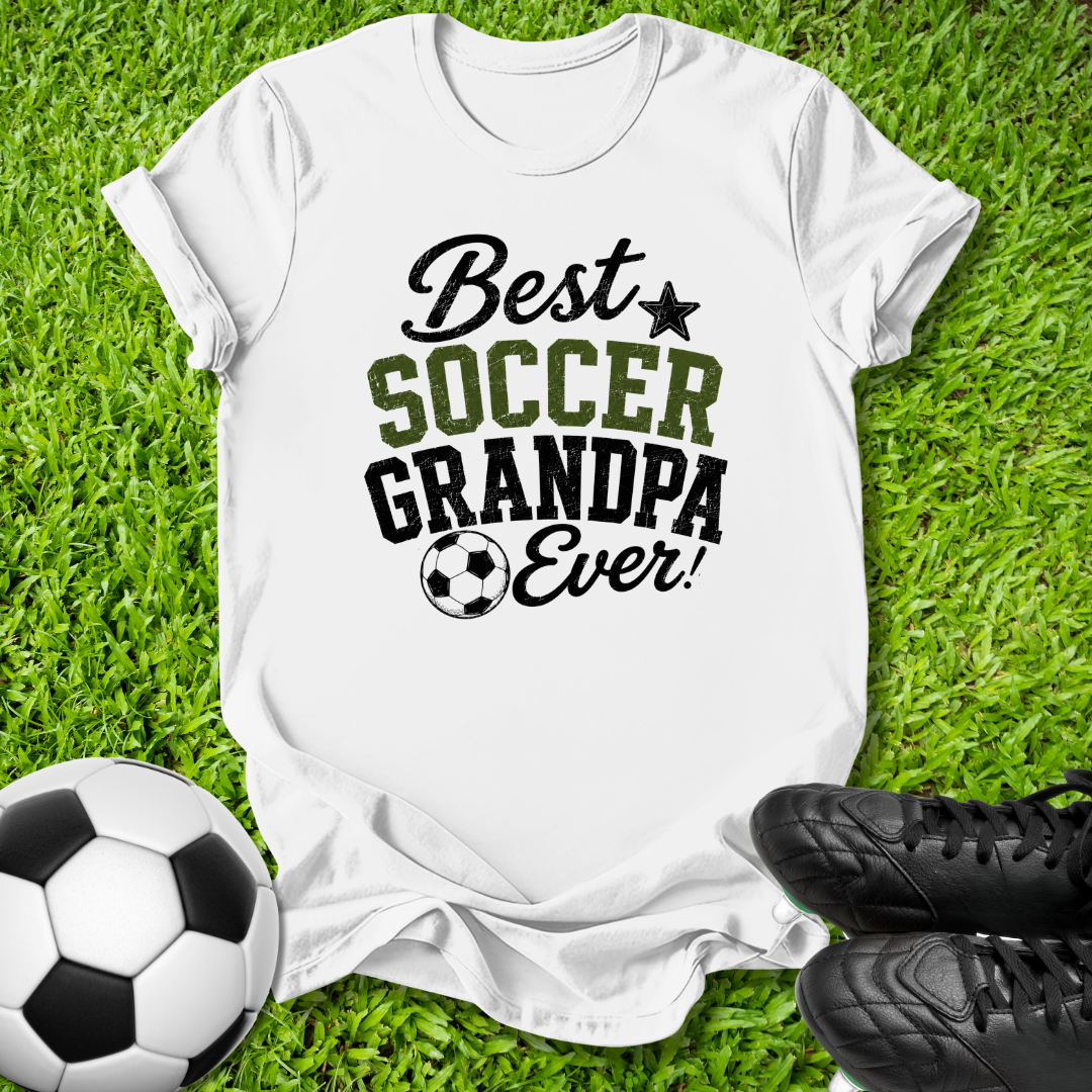 A white short-sleeve graphic T-shirt laid flat on green grass, with a soccer ball at the lower left and black cleats at the lower right. The shirt reads 'Best SOCCER GRANDPA Ever!' in black and olive-green lettering with a small soccer ball graphic.