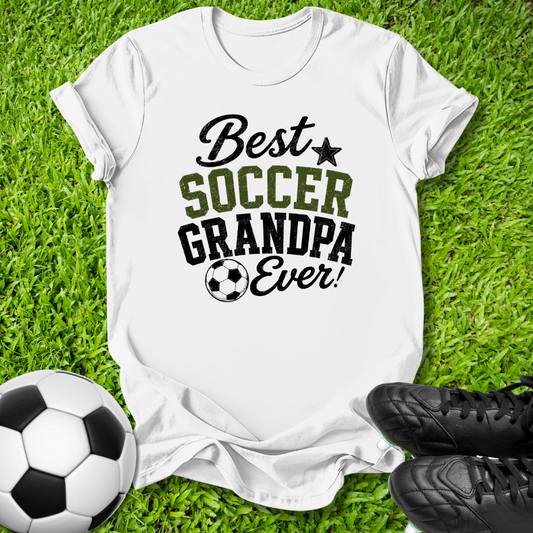 A white short-sleeve graphic T-shirt laid flat on green grass, with a soccer ball at the lower left and black cleats at the lower right. The shirt reads 'Best SOCCER GRANDPA Ever!' in black and olive-green lettering with a small soccer ball graphic.