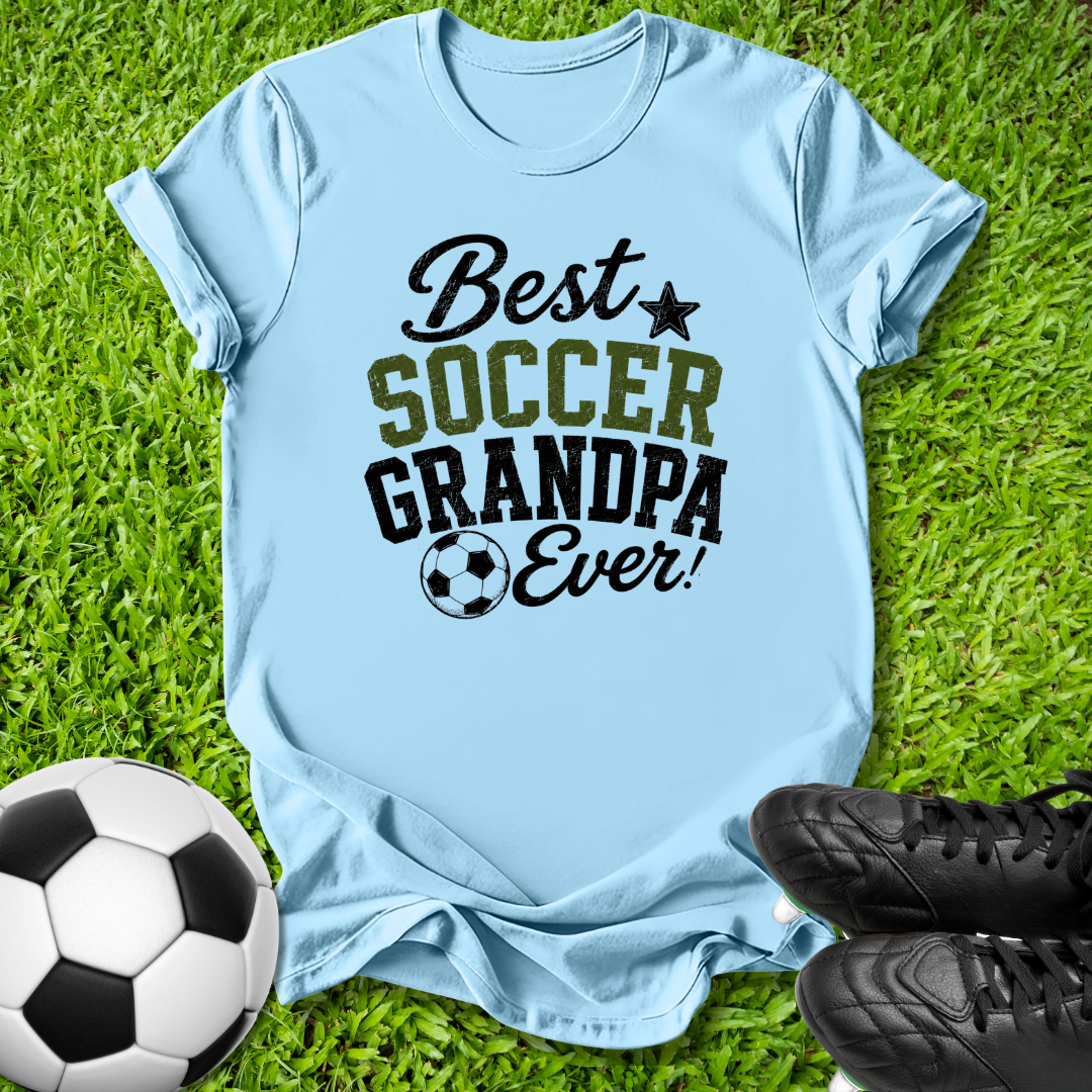 A light blue graphic t-shirt laid flat on green grass, featuring the text 'Best SOCCER GRANDPA Ever!' in black and olive green lettering. A soccer ball is on the bottom left, and a pair of black soccer cleats is on the bottom right.