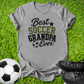 A heather gray graphic t-shirt laid flat on green grass, featuring the text 'Best SOCCER GRANDPA Ever!' in black and olive green. A soccer ball is on the left, and a pair of black cleats is on the bottom right.