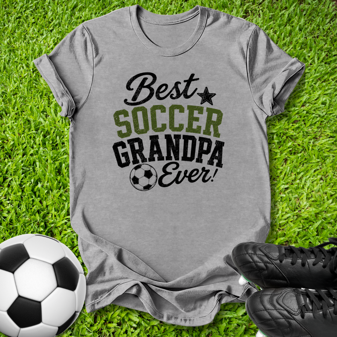 A heather gray graphic t-shirt laid flat on green grass, featuring the text 'Best SOCCER GRANDPA Ever!' in black and olive green. A soccer ball is on the left, and a pair of black cleats is on the bottom right.