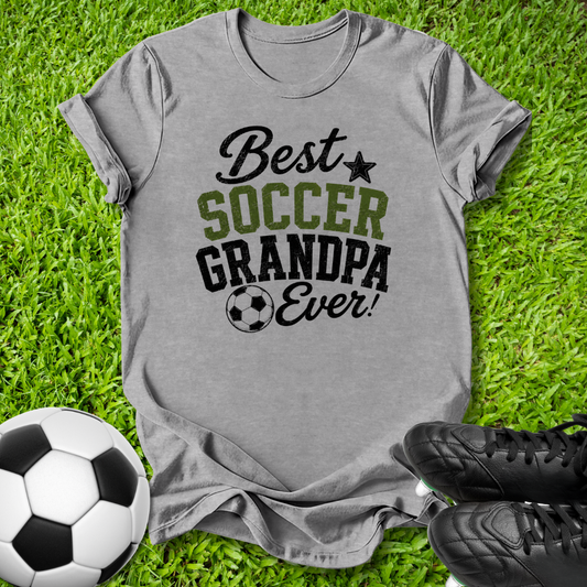 A heather gray graphic t-shirt laid flat on green grass, featuring the text 'Best SOCCER GRANDPA Ever!' in black and olive green. A soccer ball is on the left, and a pair of black cleats is on the bottom right.
