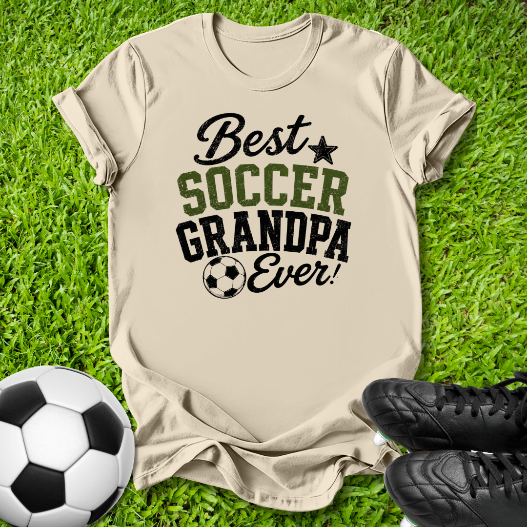 A beige graphic t-shirt laid flat on green grass, featuring the text 'Best SOCCER GRANDPA Ever!' in black and green lettering. A soccer ball is on the left, and a pair of black cleats is on the right, creating a themed flat-lay composition.