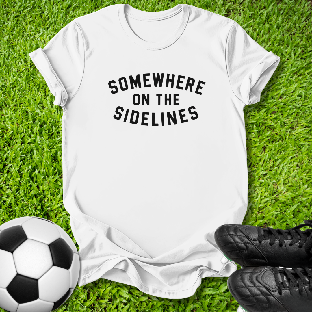 A white short-sleeve graphic T-shirt laid flat on green grass, with black text reading 'SOMEWHERE ON THE SIDELINES.' A soccer ball appears at the lower left and black cleats at the lower right, suggesting a sports-themed flat lay.