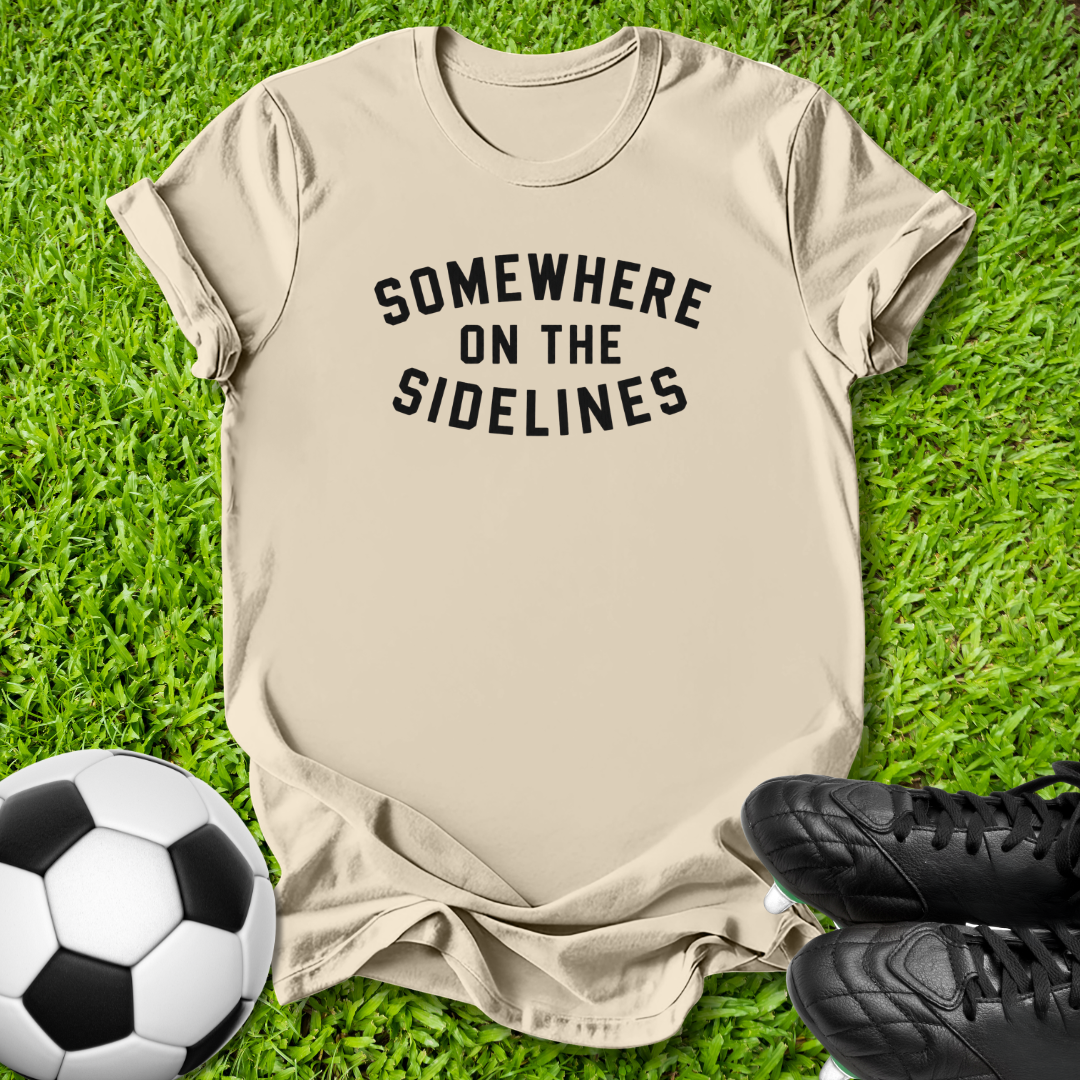 A beige graphic t-shirt with the text 'SOMEWHERE ON THE SIDELINES' printed in black arched letters, laid flat on green grass with a soccer ball and black cleats as props.