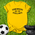 A bright yellow t-shirt with the black text 'SOMEWHERE ON THE SIDELINES' printed across the chest, laid flat on green grass with a soccer ball and black cleats as props.