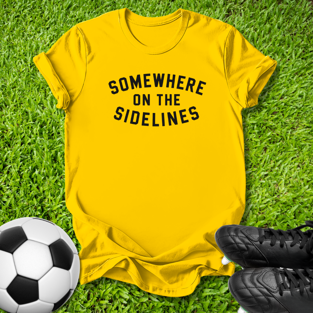 A bright yellow t-shirt with the black text 'SOMEWHERE ON THE SIDELINES' printed across the chest, laid flat on green grass with a soccer ball and black cleats as props.