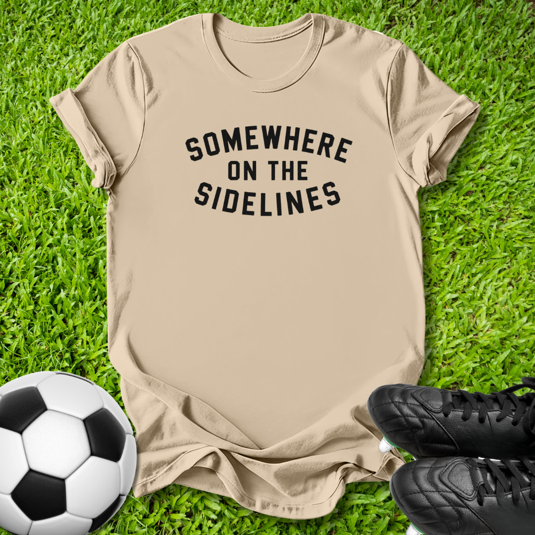 A beige short-sleeve t-shirt with the black text 'SOMEWHERE ON THE SIDELINES' printed on the chest, laid flat on green grass with a soccer ball and black cleats as props.