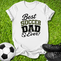 A white short-sleeve T-shirt laid flat on green grass, printed with the phrase 'Best Soccer Dad Ever!' in black and olive lettering, with a soccer ball graphic. A soccer ball and black cleats are placed around it as props.