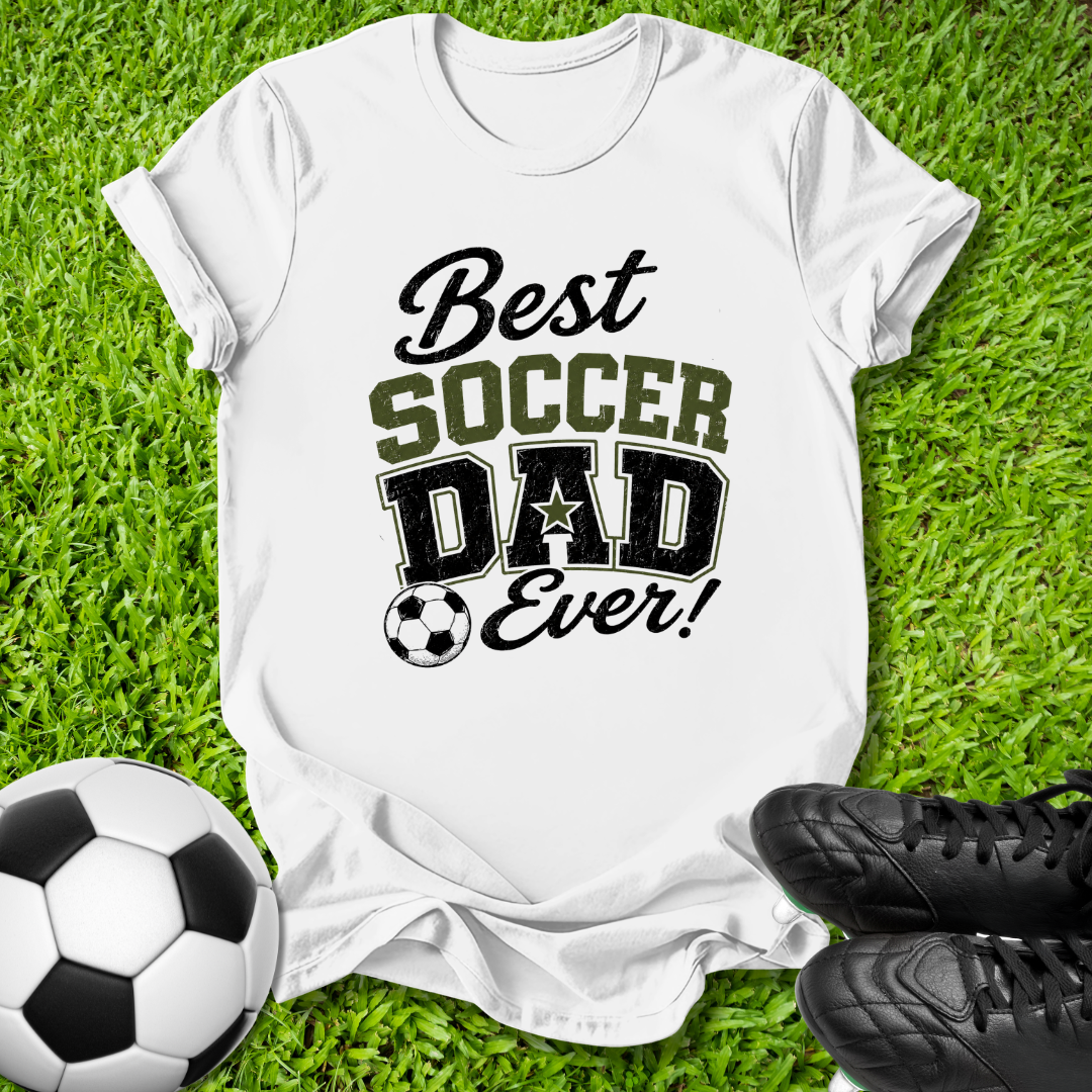 A white short-sleeve T-shirt laid flat on green grass, printed with the phrase 'Best Soccer Dad Ever!' in black and olive lettering, with a soccer ball graphic. A soccer ball and black cleats are placed around it as props.
