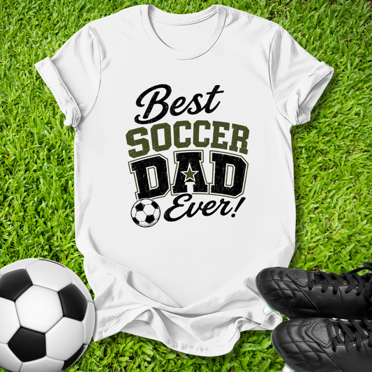 A white short-sleeve T-shirt laid flat on green grass, printed with the phrase 'Best Soccer Dad Ever!' in black and olive lettering, with a soccer ball graphic. A soccer ball and black cleats are placed around it as props.