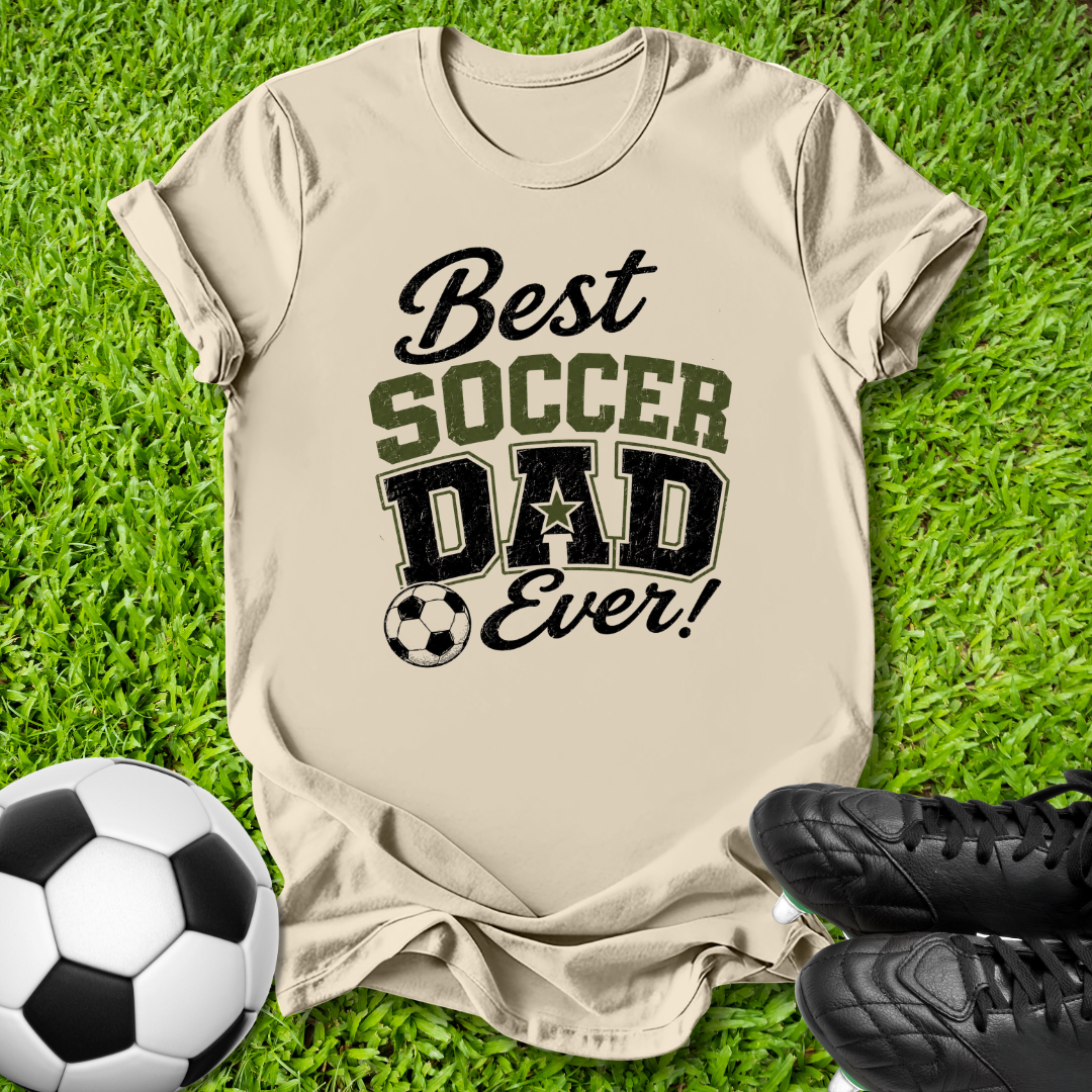 A beige graphic t-shirt laid flat on green grass, featuring the text 'Best SOCCER DAD Ever!' in black and olive green lettering, styled with a soccer ball and black cleats.