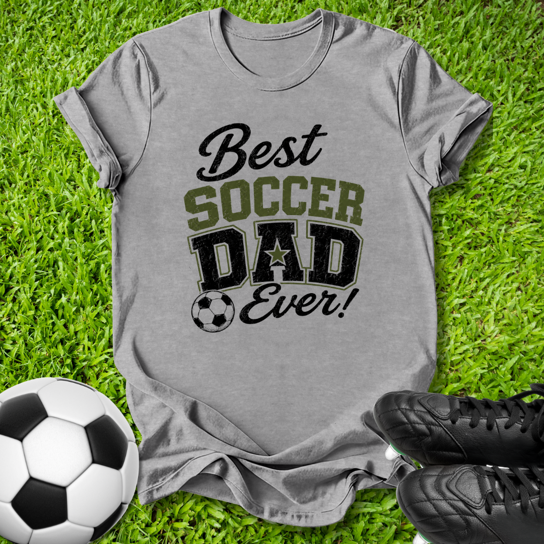 A heather gray graphic t-shirt laid flat on green grass, featuring the text 'Best SOCCER DAD Ever!' in black and olive green. A soccer ball is on the left, and a pair of black cleats is on the bottom right.