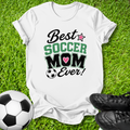A white graphic t-shirt laid flat on green grass, featuring the text 'Best SOCCER MOM Ever!' with a pink heart and soccer ball graphic. A soccer ball and black cleats are placed around it as props.