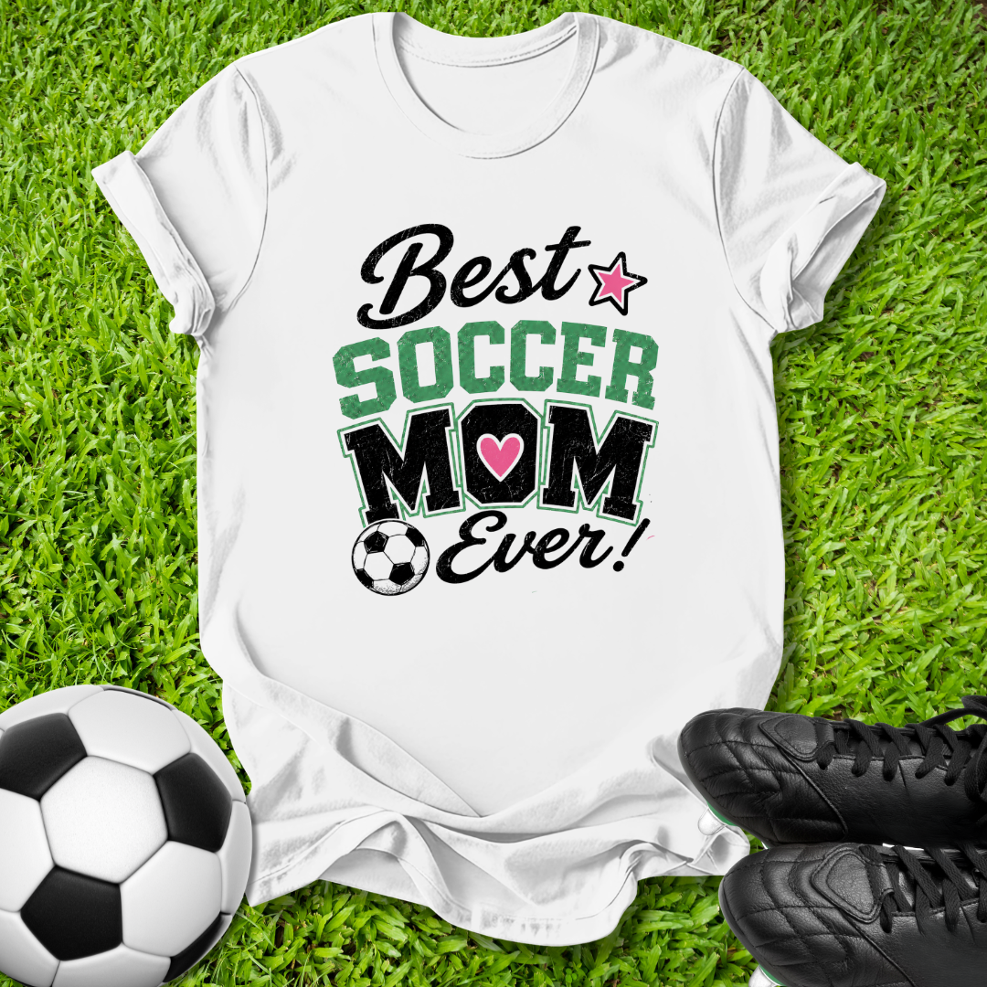 A white graphic t-shirt laid flat on green grass, featuring the text 'Best SOCCER MOM Ever!' with a pink heart and soccer ball graphic. A soccer ball and black cleats are placed around it as props.