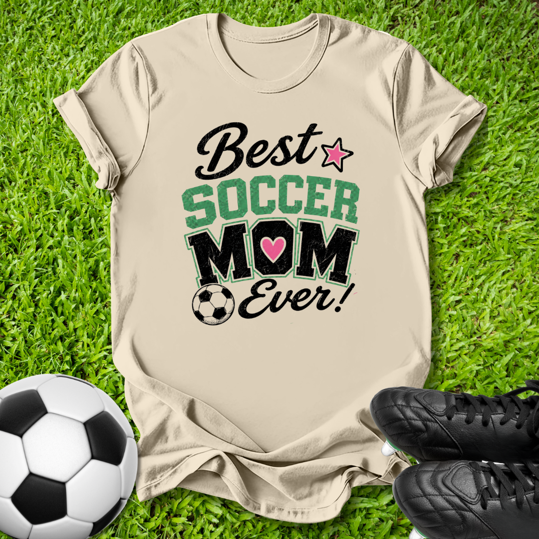 A beige graphic t-shirt laid flat on green grass, featuring the text 'Best SOCCER MOM Ever!' with a pink heart and soccer ball graphic. A soccer ball and black cleats are placed around it as props.
