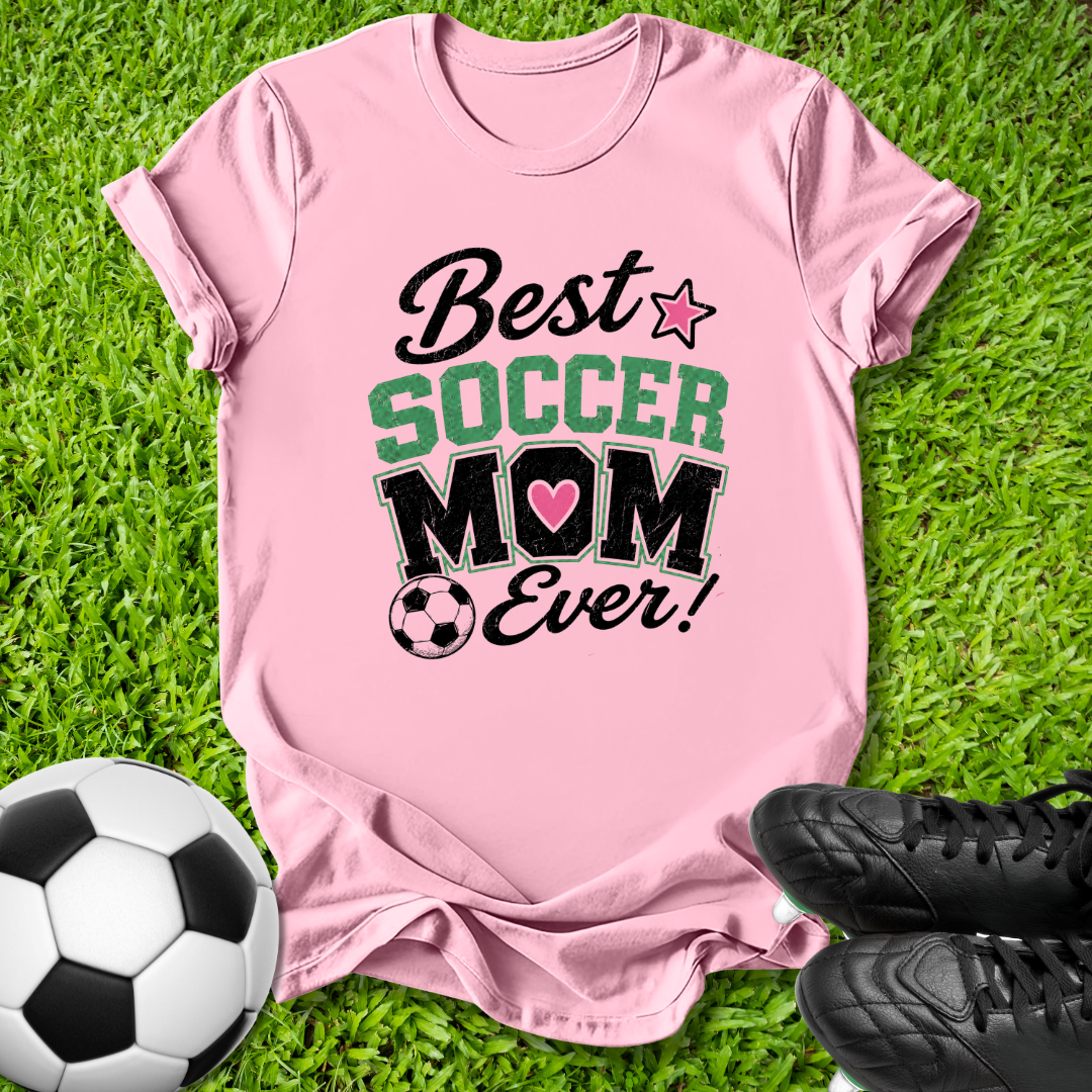 A pink short-sleeve t-shirt laid flat on green grass with a soccer ball and black cleats nearby. The shirt has a graphic that reads 'Best SOCCER MOM Ever!' with a pink star, green 'SOCCER' text, and a pink heart inside the 'O'.