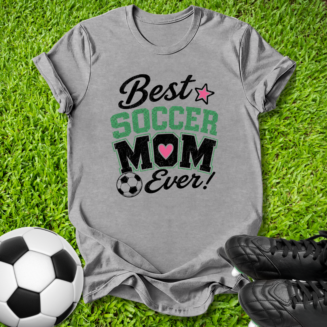 A heather gray graphic t-shirt laid flat on green grass, featuring the text 'Best SOCCER MOM Ever!' with a pink heart and star. A soccer ball is on the left and black cleats are on the right.