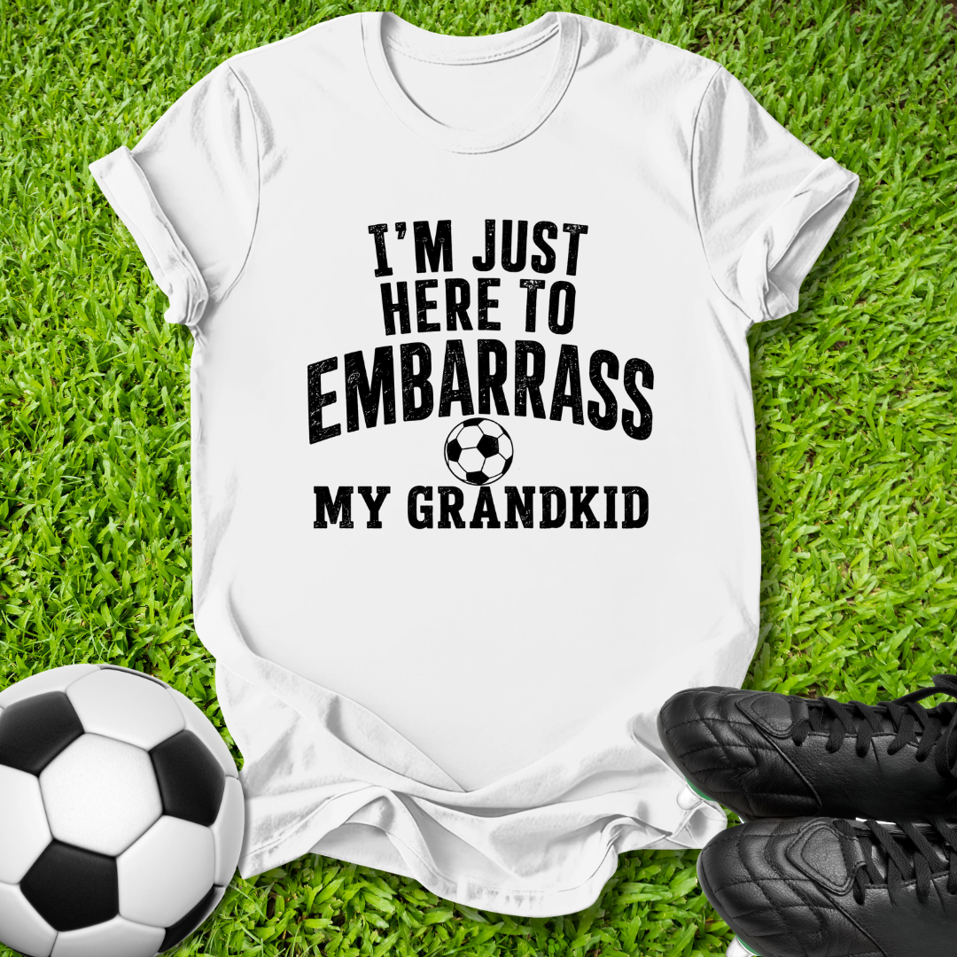 A white short-sleeve graphic T-shirt laid flat on green grass, with a soccer ball on the left and black cleats on the right. The shirt has bold black text reading “I'M JUST HERE TO EMBARRASS MY GRANDKID” with a small soccer ball graphic in the center.