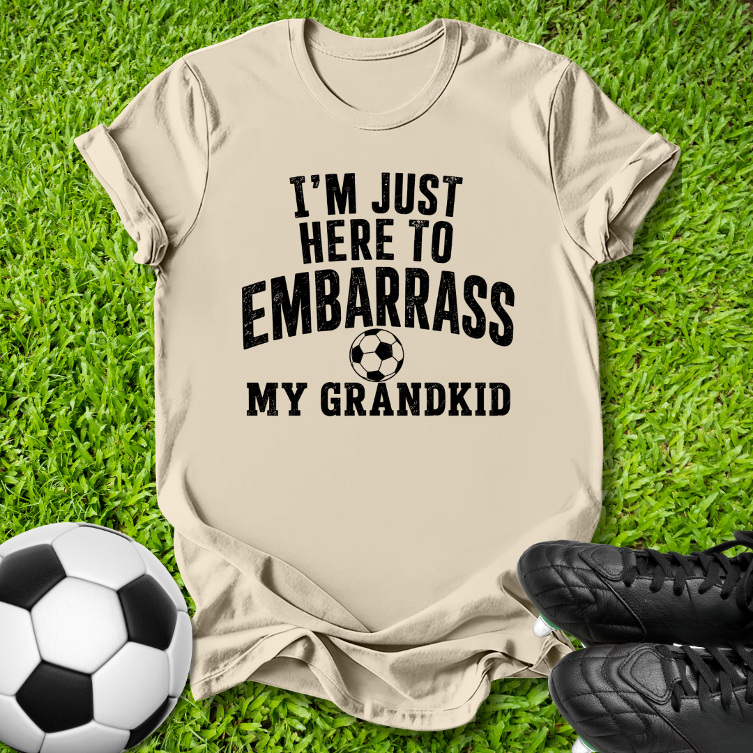 A beige graphic t-shirt laid flat on green grass, featuring the text 'I'M JUST HERE TO EMBARRASS MY GRANDKID' in black with a soccer ball graphic. A black-and-white soccer ball and black cleats are placed around it as props.