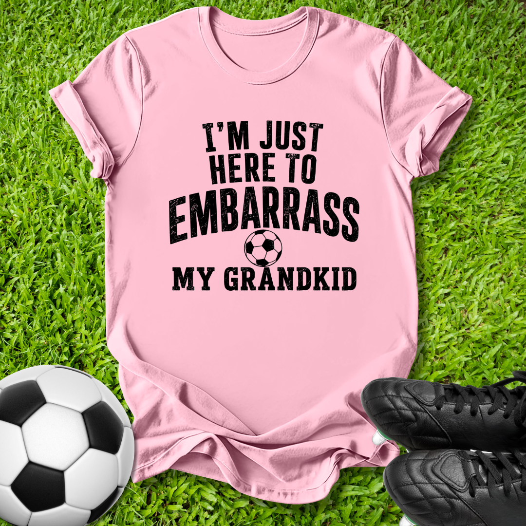 A pink short-sleeve graphic t-shirt laid flat on green grass, with a soccer ball on the left and black cleats on the right. The shirt features the black printed text 'I'M JUST HERE TO EMBARRASS MY GRANDKID' with a small soccer ball graphic in the center.