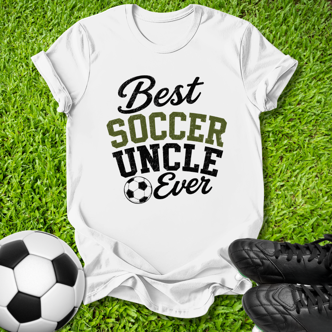 A white graphic t-shirt laid flat on green grass, featuring the text 'Best SOCCER UNCLE Ever' in black and olive green lettering with a soccer ball icon. A soccer ball is on the left and black cleats are on the right.
