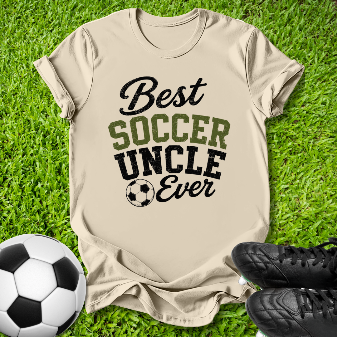 A beige graphic t-shirt laid flat on green grass, featuring the text 'Best SOCCER UNCLE Ever' in black and olive green lettering with a soccer ball icon. A black-and-white soccer ball and black cleats are placed around it as props.