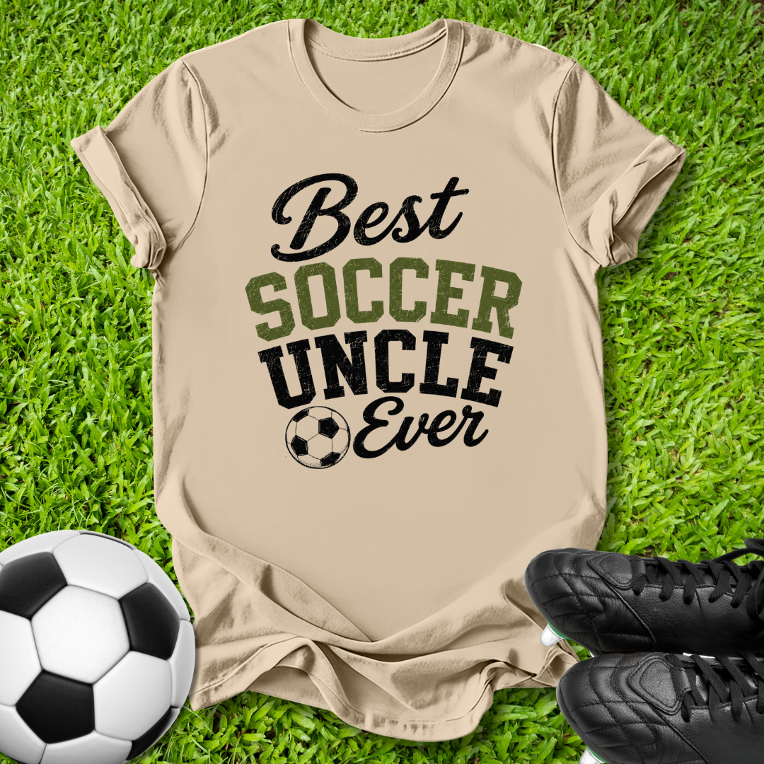 A beige graphic t-shirt laid flat on green grass, featuring the text 'Best SOCCER UNCLE Ever' in black and olive green lettering with a soccer ball icon. A soccer ball and black cleats are placed around it as props.