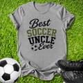 A heather gray graphic t-shirt laid flat on green grass, featuring the text 'Best SOCCER UNCLE Ever' in black and olive green lettering with a soccer ball icon. A black-and-white soccer ball and black cleats are placed around it as props.