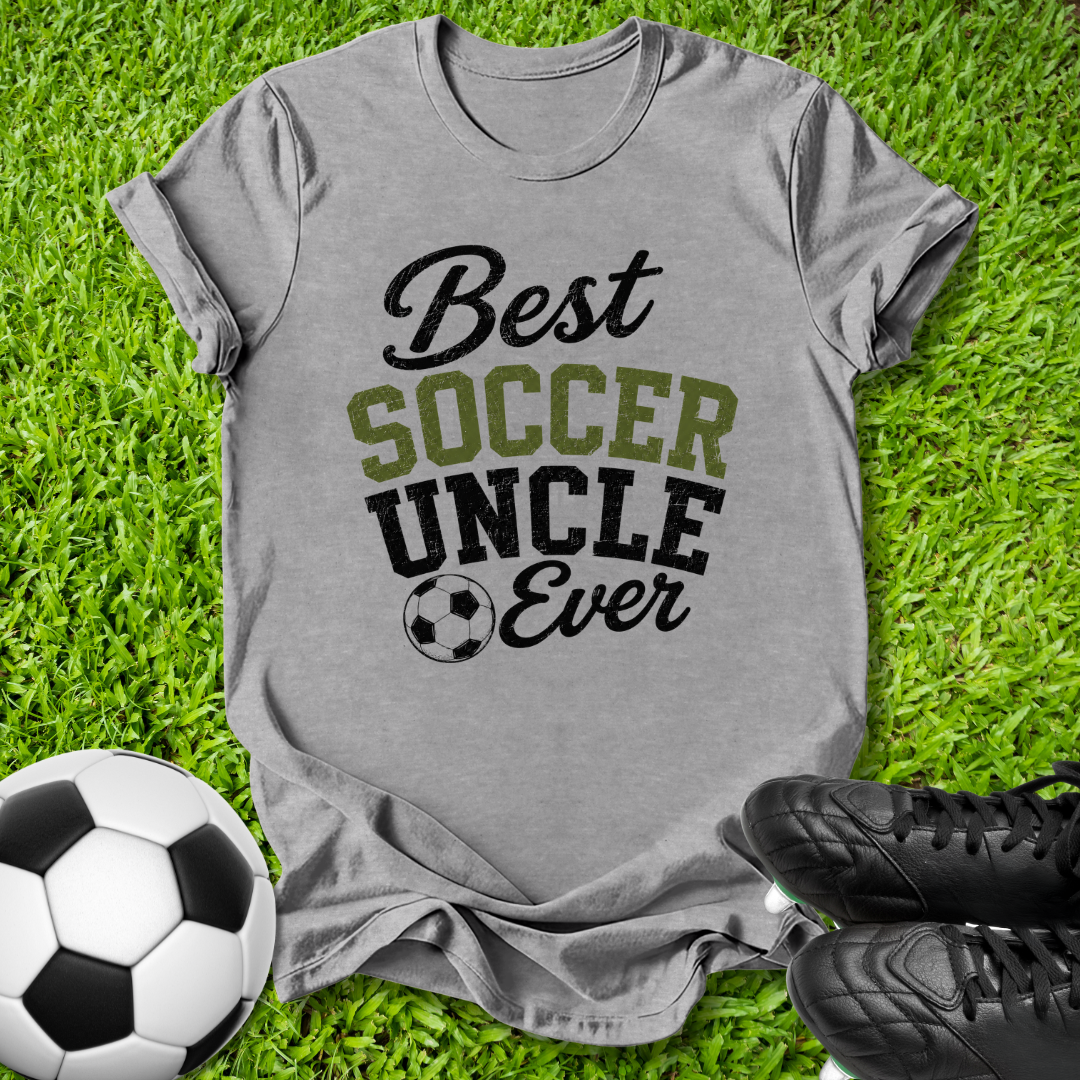 A heather gray graphic t-shirt laid flat on green grass, featuring the text 'Best SOCCER UNCLE Ever' in black and olive green lettering with a soccer ball icon. A black-and-white soccer ball and black cleats are placed around it as props.