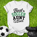 A white graphic t-shirt laid flat on green grass, featuring the text 'Best SOCCER AUNT Ever' in black and green lettering with a small soccer ball icon. A black-and-white soccer ball is on the left, and a pair of black cleats is on the right, creating a soccer-themed flat lay.