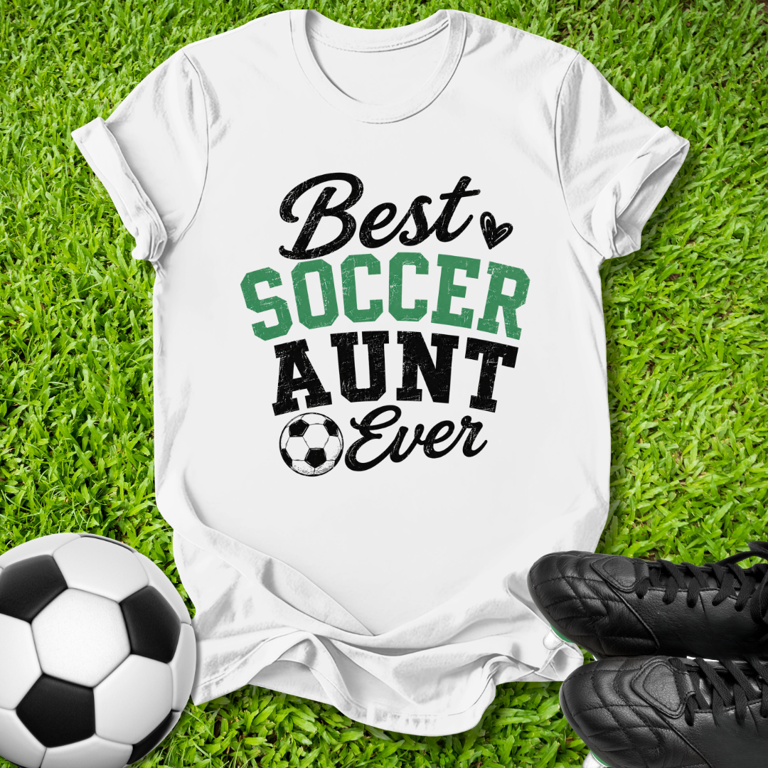 A white graphic t-shirt laid flat on green grass, featuring the text 'Best SOCCER AUNT Ever' in black and green lettering with a small soccer ball icon. A black-and-white soccer ball is on the left, and a pair of black cleats is on the right, creating a soccer-themed flat lay.