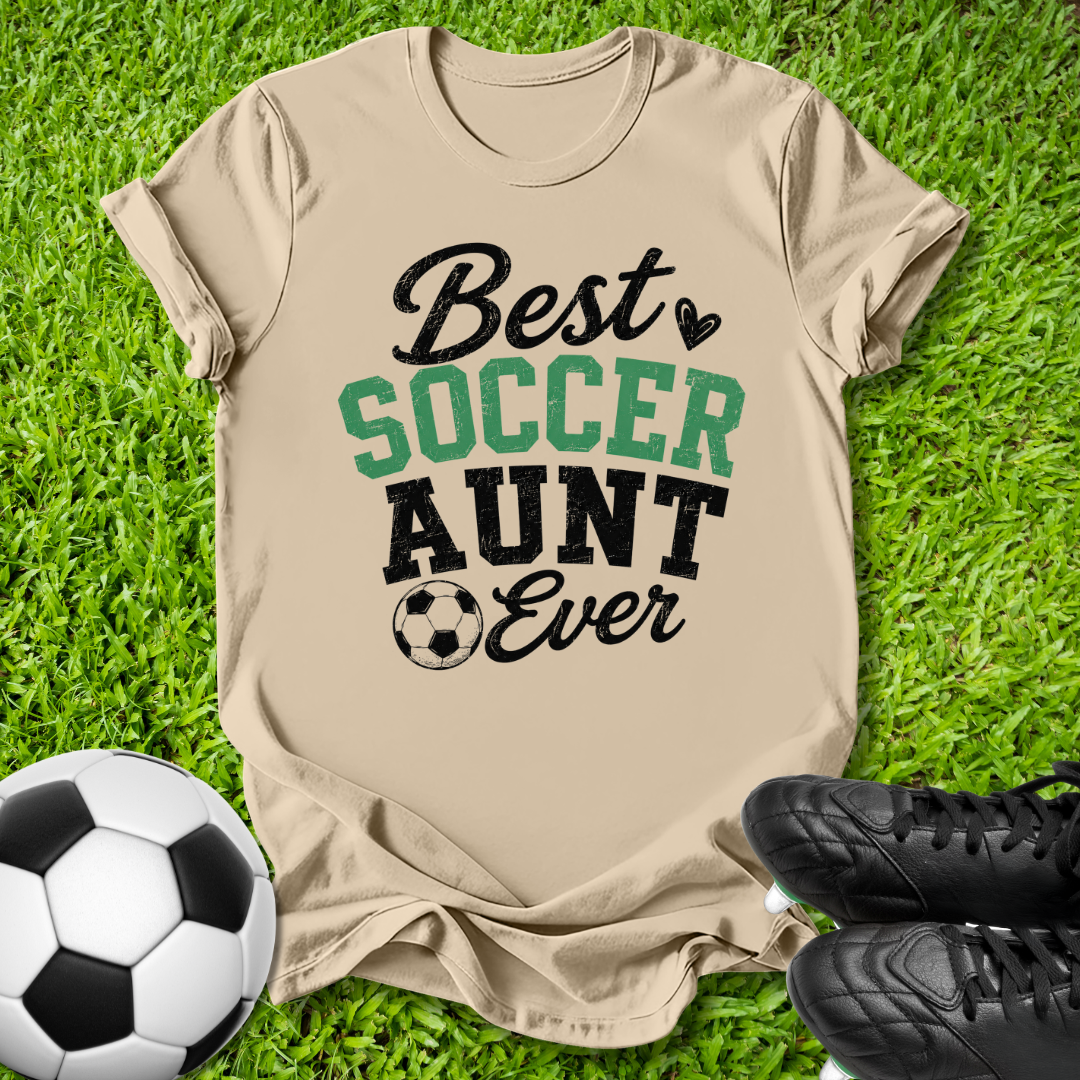 A beige graphic t-shirt laid flat on green grass, featuring the text 'Best SOCCER AUNT Ever' in black and green lettering with a small soccer ball icon. A black-and-white soccer ball is on the left, and a pair of black cleats is on the right, creating a themed flat-lay composition.
