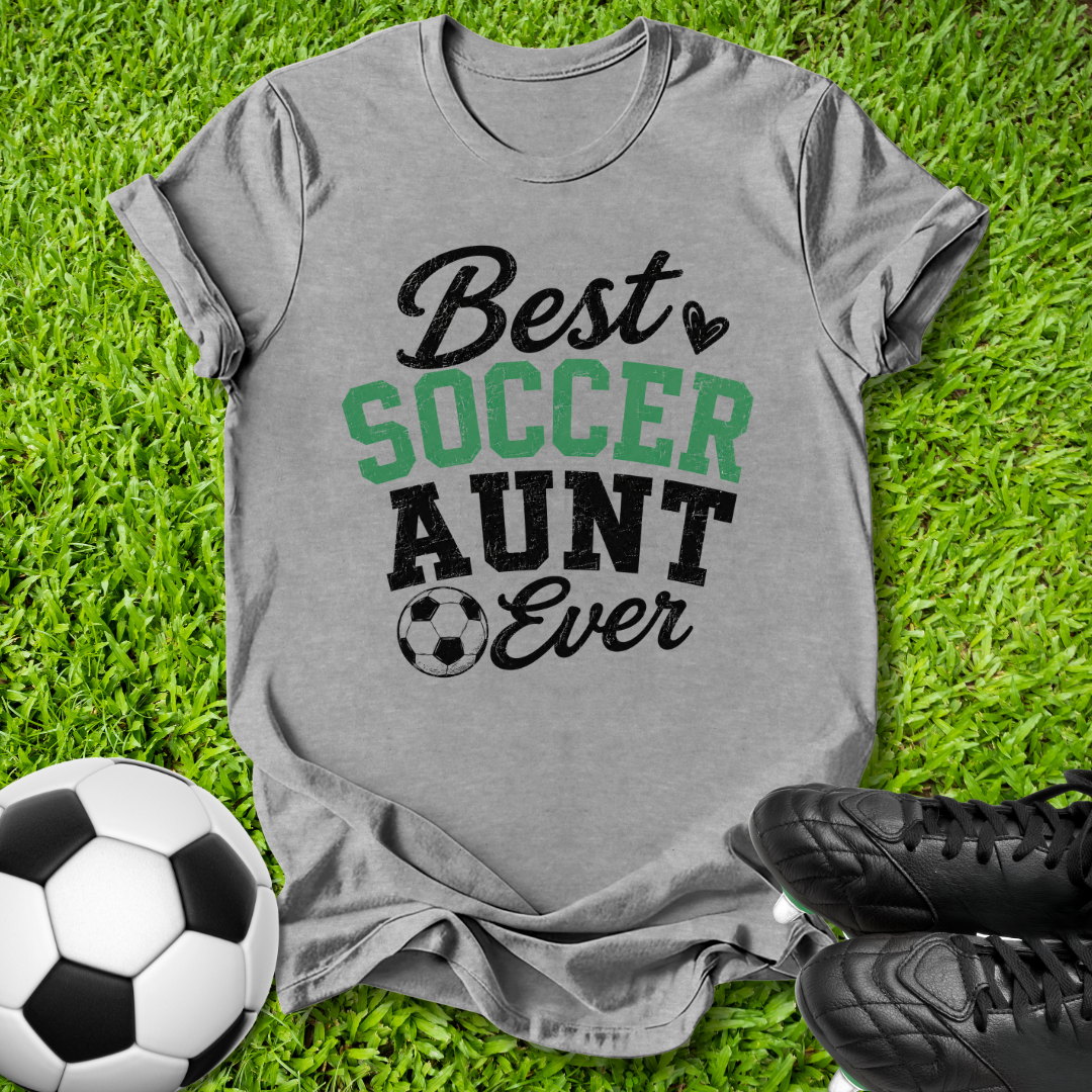 A heather gray graphic t-shirt laid flat on green grass, featuring the text 'Best SOCCER AUNT Ever' in black and green. A soccer ball is on the left, and black cleats are on the right, creating a themed flat-lay composition.