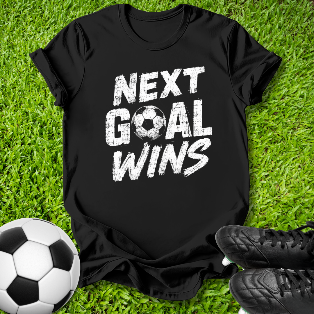 A black short-sleeve t-shirt with a distressed white graphic reading 'NEXT GOAL WINS' is laid flat on green grass, styled with a soccer ball on the left and black cleats on the right.