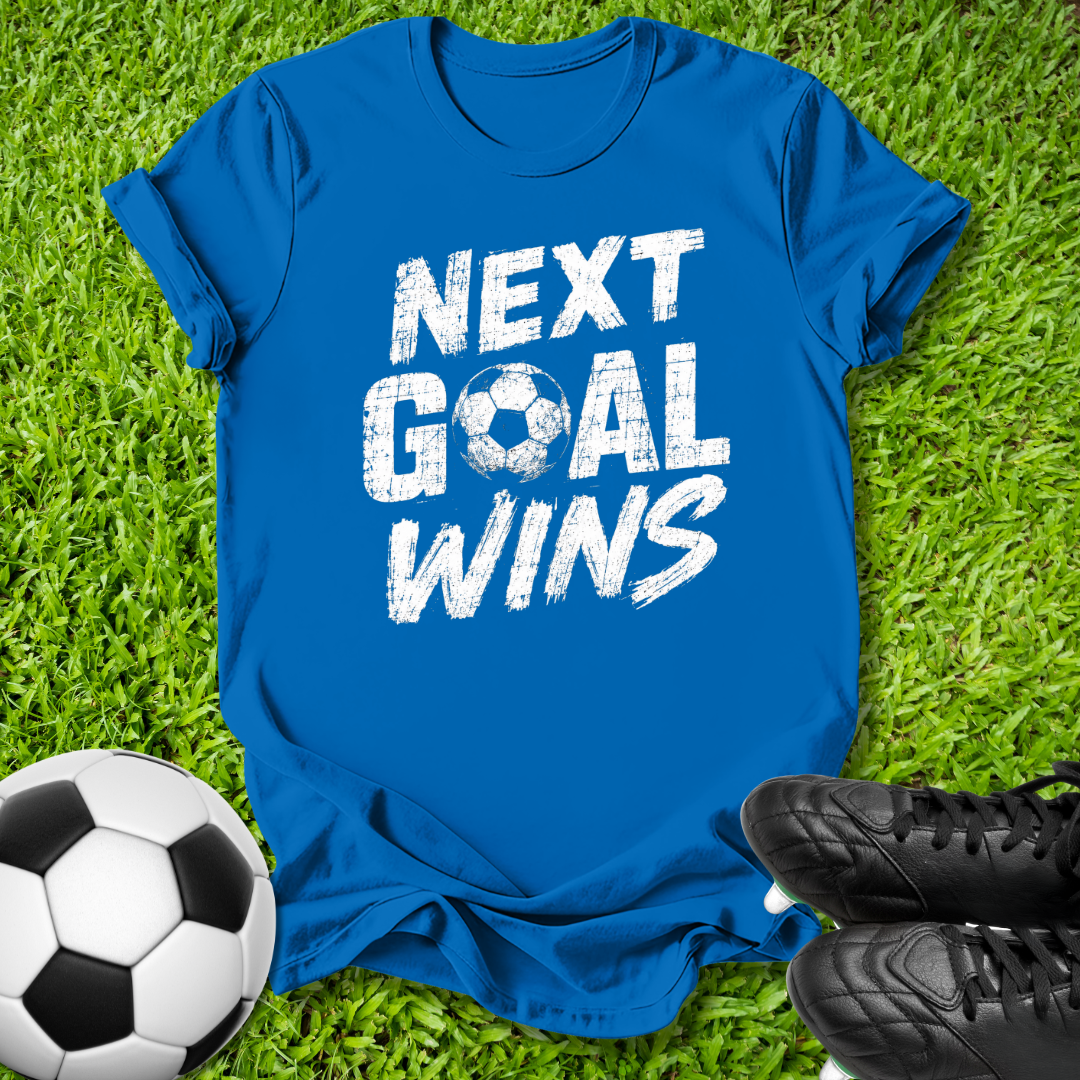 A royal blue short-sleeve t-shirt with the phrase 'NEXT GOAL WINS' printed in distressed white letters across the front, laid flat on green grass with a soccer ball and black cleats nearby.