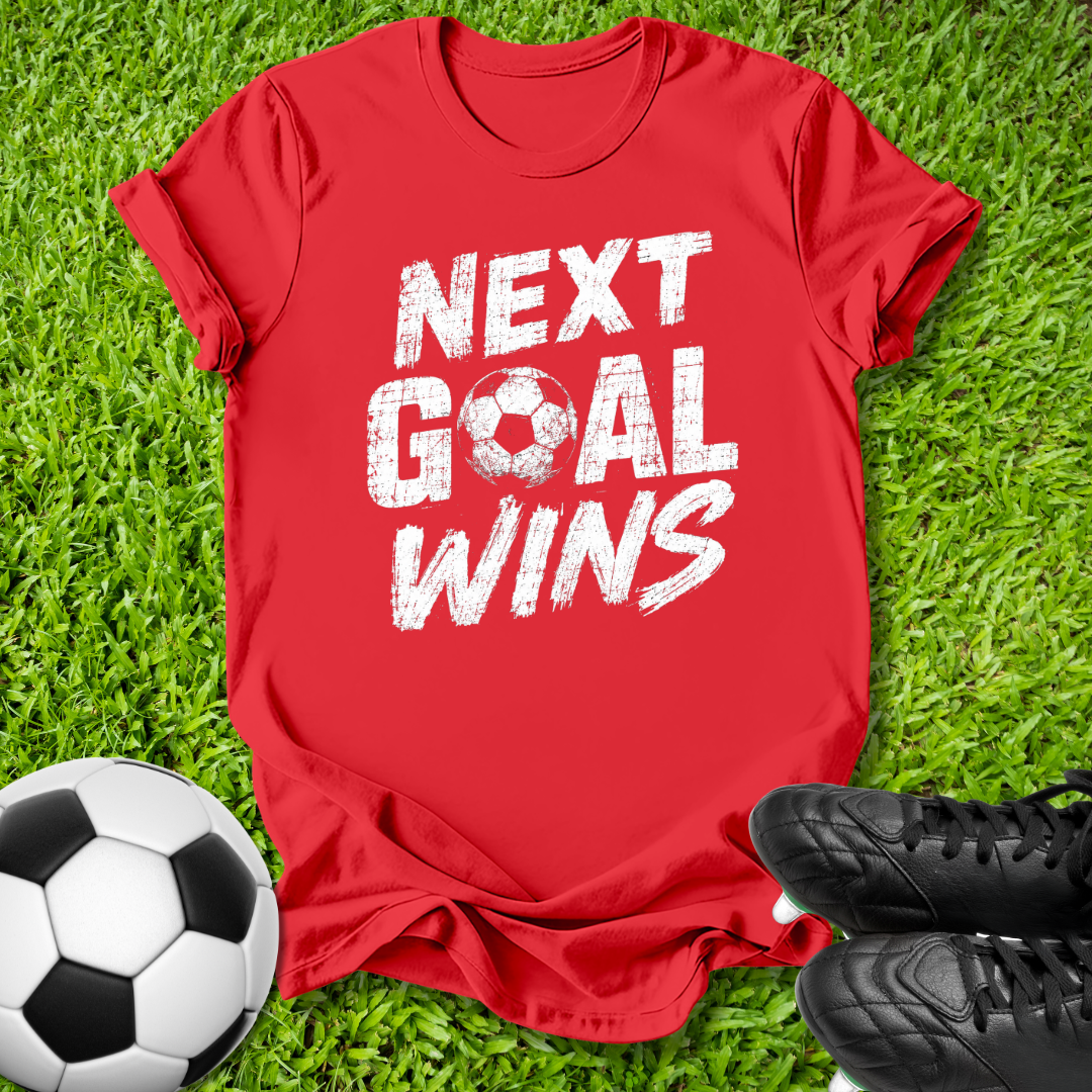 A red short-sleeve graphic T-shirt laid flat on green grass, with a soccer ball at the lower left and black cleats at the lower right. The shirt has large distressed white text reading 'NEXT GOAL WINS' with a soccer ball icon replacing the letter O.