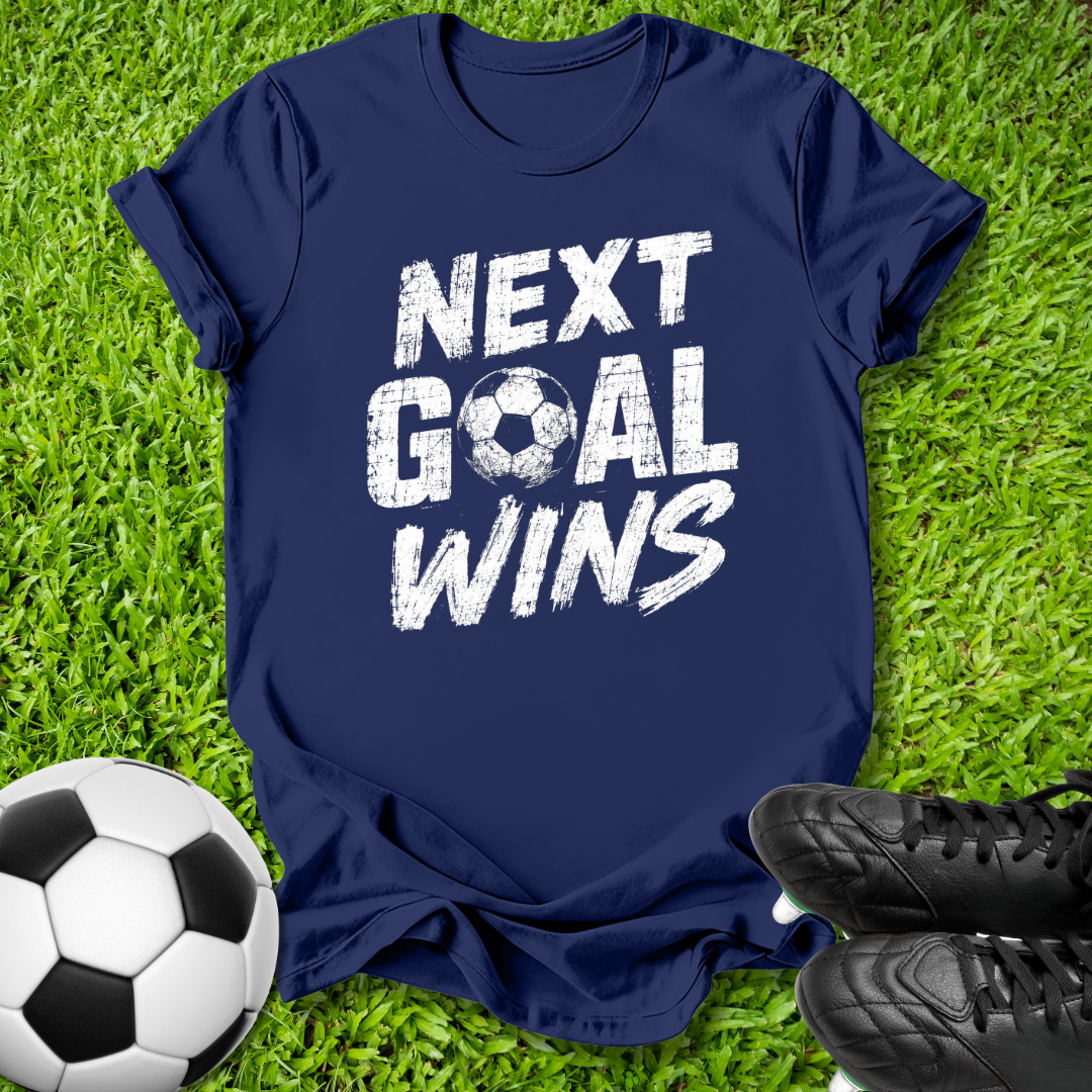 A navy blue short-sleeve t-shirt with the phrase 'NEXT GOAL WINS' printed in distressed white letters, laid flat on green grass with a soccer ball and black cleats nearby.