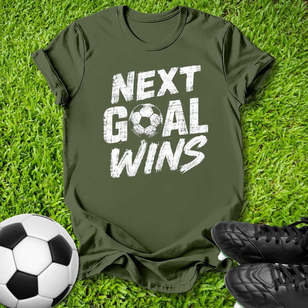 An olive green t-shirt with the text 'NEXT GOAL WINS' in distressed white lettering, laid flat on green grass with a soccer ball and black cleats as props.